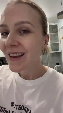 The video shows a woman with blonde hair pulled back into a small bun. She is wearing a white t-shirt with black text in Russian. She has silver hoop earrings and a ring piercing in her ear. The background shows white kitchen cabinets, a stainless steel kettle, and items on shelves. The woman is singing and talking to the camera.