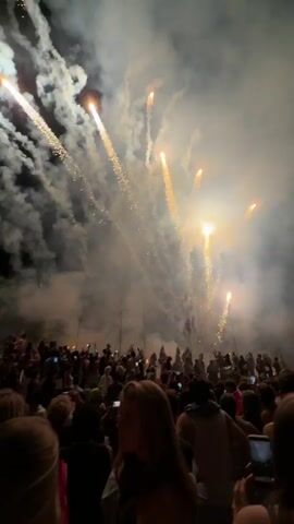 This video shows a nighttime outdoor event with fireworks. A large crowd of people, viewed from behind, are gathered to watch the display. The sky is filled with smoke and multiple bursts of fireworks, primarily in shades of yellow, orange, and white, with a segment of bright red fireworks towards the end. Many individuals in the crowd are holding up their phones to record the event. The overall atmosphere appears festive and celebratory.