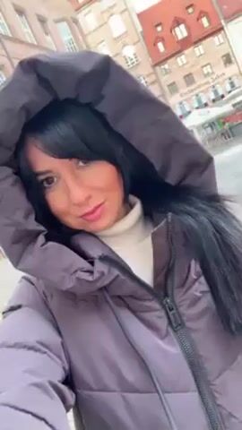 A woman stands outside in a town square on an overcast day. The woman wears a puffy winter coat with a large hood up, a cream colored turtleneck, and has long black hair with bangs. She faces the camera with a slight smile. Behind her are buildings of various sizes. Some of the buildings have red tile roofs. There are some partially visible vendor booths in the background. A statue is partially visible on the right of the screen.