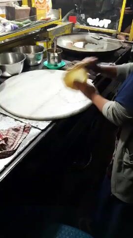 The video shows a person's hands working on a circular, white surface, likely a marble slab, on what appears to be a food stall or kitchen counter. The person is rolling out a thin, circular piece of dough, shaping it with their hands. Behind the marble slab, there are several stainless steel bowls and a container with a green lid. Further in the background, a large, flat griddle or pan is visible, suggesting food preparation is underway. The environment seems to be a bustling, possibly outdoor, setting, indicated by the general lighting and background activity. The person is wearing a blue garment. The focus is on the skillful manipulation of the dough.