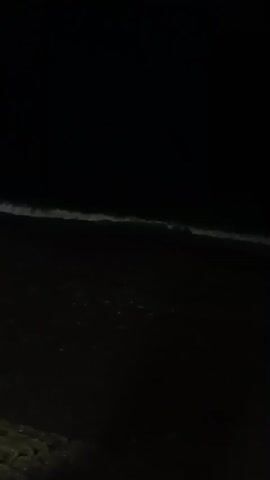 The video is a dark, grainy, still shot of a beach at night. Small waves are breaking on the shore, and some distant lights are visible on the horizon. The foreground shows a textured surface that appears to be sand or gravel.