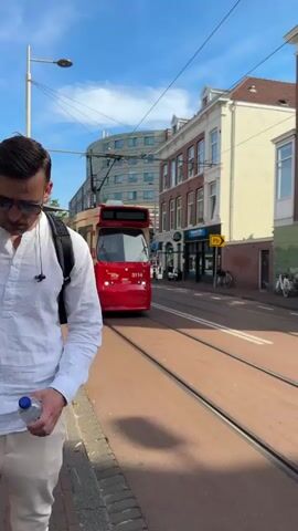 A red and white tram with the number 3114 is traveling down a street. The tram has its doors open. The street is paved with red brick. Buildings with windows are visible on the right side of the street. A person wearing a white shirt and beige pants is standing on the sidewalk on the left side of the street. The sky is blue with some clouds.