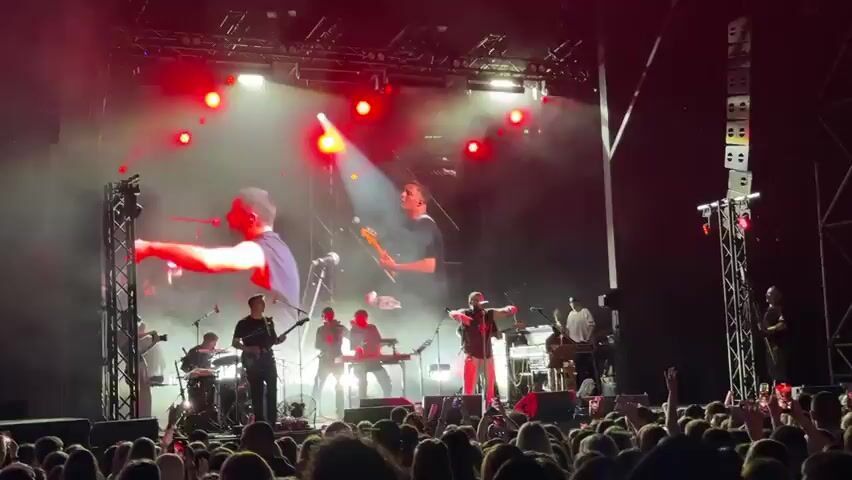 A live concert performance is depicted. A band is on stage, with a large screen behind them displaying visuals. The band members are playing instruments, including guitars, keyboards, and drums. One band member, likely the lead singer, is at the front of the stage, interacting with the audience. The audience is in front of the stage, with many people holding up their phones to record the performance. The stage is lit with spotlights and stage lighting, creating a dynamic atmosphere. The screen behind the band displays the name "NAVAI". The overall scene suggests a high-energy musical event.