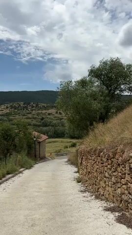 A dirt road curves through a rural landscape under a cloudy sky. On the right, a rough stone wall runs alongside the road, bordered by dry grass. On the left, a small, stone building with a red-tiled roof is partially visible, with greenery and a lamppost next to it. The road leads into the distance towards a valley filled with trees and rolling hills covered in vegetation. The sky is partly cloudy, with patches of blue visible between the white clouds.