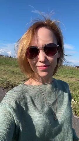 This is a selfie video of a woman with short, reddish-blonde hair and sunglasses. She is wearing a light green, knitted sweater with a wide neckline and a delicate gold necklace. She is outdoors, standing on an asphalt path with a grassy area and a fence behind her. In the distance, there are houses and hills under a bright blue sky with some clouds. The sun is shining, suggesting it is daytime. The woman is smiling and looking at the camera, appearing relaxed and happy. The scene suggests a casual, perhaps rural or suburban setting in Mursalimkino, Russia.