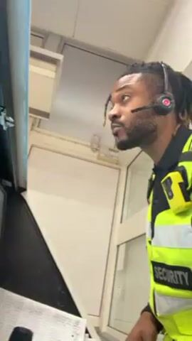 Here's a detailed description of the video:

A man is the subject of the video, wearing a bright yellow security vest with "SECURITY" printed on it. He has dreadlocks pulled back in a half ponytail style and is wearing a headset with a microphone. He has a dark complexion, a trimmed beard, and a piercing in his left nostril. 

The man is positioned inside what appears to be a control room or office. He is standing near a desk with paperwork and appears to be looking at computer screens that are slightly out of view. He gestures with his hands as he speaks, looking directly into the camera at times.

The environment is indoors. The room appears to be well-lit, with visible components of an air conditioning or ventilation system. The walls are white. There are window panes visible to the right.