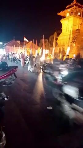 The video, filmed at night, shows a street scene with several people and motorcycles. Indonesian flags are visible, suggesting a possible celebration or event. A prominent, illuminated building with a clock tower stands in the background. Some individuals are walking, while others are on or near motorcycles. The lighting is dim, with artificial lights illuminating the scene. Motion blur suggests movement within the frame, particularly with the motorcycles. The ground appears to be paved.