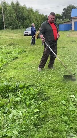 A man wearing a red shirt, green jacket, and camouflage pants is using a string trimmer to cut tall grass and weeds. He is standing in a grassy yard with trees in the background. There is a car parked in the distance and a building with a blue roof. The trimmer is creating a cloud of cut grass. The video is shot from a low angle, looking up at the man.