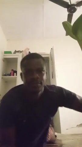 This media file captures a short, low-angle video of a person performing push-ups indoors. The primary subject is a Black male, seen from the chest up, actively engaged in the exercise. He is wearing a dark t-shirt and appears to be on a wooden floor. His face is somewhat obscured by motion blur during the exertion of the push-ups.

The setting appears to be a room within a residence. To the left of the frame, there is a white cabinet with its door ajar, revealing shelves with various items, including what look like bottles and containers. Above the cabinet, there are some objects placed on a shelf. In the background, a white closet or door is visible. A ceiling fan with four blades is mounted on the ceiling, and a plant with large green leaves is positioned in the upper right corner of the frame. The lighting suggests it is daytime, possibly late morning or afternoon, given the ambient light. There is no visible text within the media file. The activity is clearly physical exertion, specifically push-ups, and the overall mood is one of exercise and focus. There are no strong indications of weather or specific time of day beyond the general indoor lighting. The location context of Abeokuta, Nigeria, does not offer any specific visual cues within the frame itself.