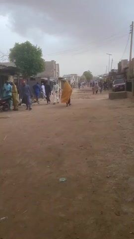 The video shows a dirt road in what appears to be a village or small town. People are walking along the road in both directions. The sky is overcast and a sandstorm is forming. Buildings line the road, and a few trees are visible. A car is parked on the right side of the road. There is trash on the ground.