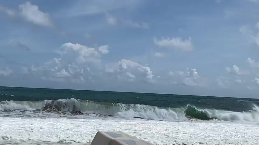 A wide shot shows a choppy ocean with large waves breaking near the shore under a cloudy blue sky. A part of a white structure, possibly a railing or a bench, is visible in the foreground. The waves are a mix of deep blue and white foam as they crash onto the beach. The sky is partly cloudy with scattered white clouds. The overall scene suggests a dynamic and somewhat rough coastal environment.