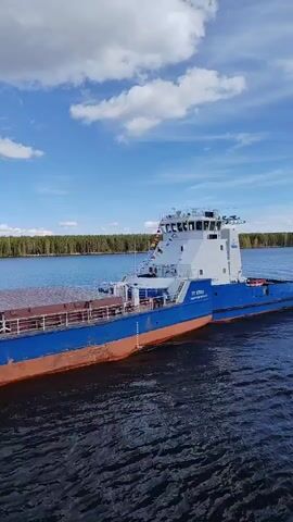A blue and white tugboat, labeled "PT 82809 Санкт-Петербург" (Saint Petersburg) and "ВЕГА" (Vega) in white lettering, moves on a waterway. The ship has an orange waterline. A forest lines the shore in the background. The sky is blue with fluffy white clouds. The water is dark with ripples. A small number of colorful flags are displayed on the boat. The boat is the main subject of the shot. The video is a medium shot, showing the boat in motion, taken from a slightly elevated position.