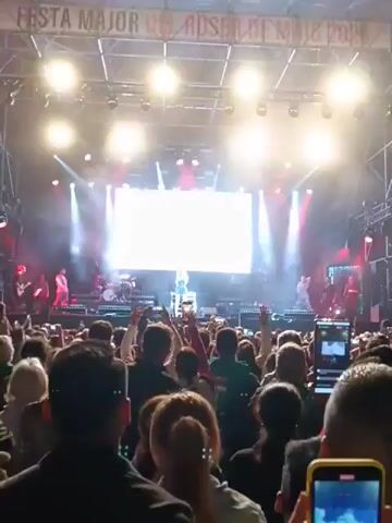 The video shows a female pop singer performing on a brightly lit stage in front of a large crowd. The singer is wearing a sparkly blue dress, and she is standing in the center of the stage, on a white box. Behind her is a large, bright white screen, flanked by red and white spotlights. Flanking the stage are two male dancers, wearing colorful outfits. A band can be seen behind them. The crowd is holding up phones and cameras, recording the performance. The overall environment suggests a vibrant and energetic concert setting.