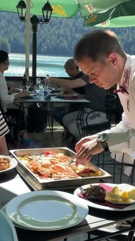 A waiter in a white shirt and red bow tie is serving food from a large silver pan to a table outdoors. The waiter is focused on serving the food. There are four people sitting at the table. In the background, there is a lake and mountains. The table is covered with a green umbrella. There are plates, cutlery, and glasses on the table.