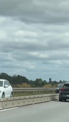 The video shows a view from a moving vehicle on a highway. In the foreground, there is a concrete barrier. Two cars are visible on the highway: a dark gray station wagon in the left lane and a black SUV in the right lane. The black SUV has yellow lines on its rear bumper and a license plate that reads "FL CUD 325". In the background, there are several semi-trucks, trees, and a cloudy sky. The vehicle appears to be driving on a multi-lane highway.