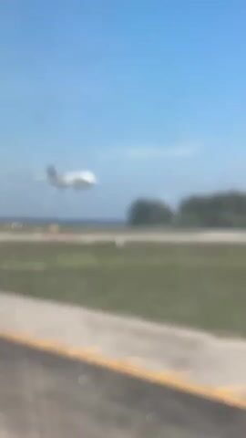 A plane, labeled "THAI," is in the air, positioned for landing at a runway surrounded by grassy fields. The sky above is clear and blue. A strip of road with yellow lines runs along the bottom left. Trees are visible beyond the runway. An automated voice message is heard.