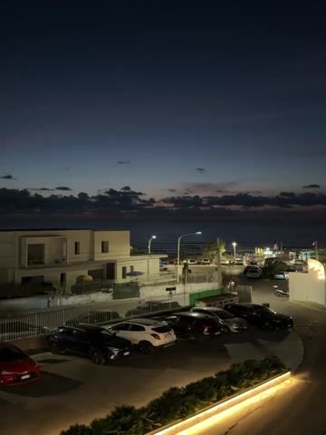 The video shows a dark blue twilight sky over a body of water, likely the sea, with clouds casting shadows. In the foreground, a parking lot is visible with several cars parked, including a red car, a blue car, a white car, and several black cars. There is a white building to the left of the parking lot, and further back, along the waterfront, there are more buildings and lights. A streetlamp is visible, and the scene appears to be near a coastal area. The lighting suggests it is dusk or early evening.