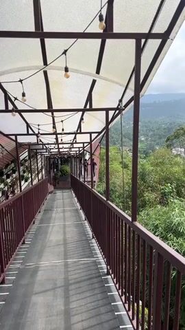 The video shows an elevated walkway with a translucent roof and a dark red-brown metal railing, overlooking a lush, mountainous landscape. The walkway has a dark surface with white markings and string lights hanging from its roof structure. In the distance, green trees and bamboo are abundant, with some scattered buildings visible among the foliage. Hazy mountains rise in the far background under a partly cloudy sky. Several indistinct figures can be seen walking on the walkway further down. The camera slowly pans from left to right, revealing more of the scenic view.