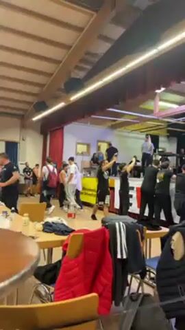The video shows a gathering in a large room with a stage. Many people are present, some are on the stage and some are in the audience area. On the stage, two people are participating in an arm-wrestling competition while others watch and record with their phones. The audience area has tables with personal items and drinks scattered around. The room has a wooden ceiling with fluorescent lights. The stage has a red curtain backdrop and banners with logos on the front. The atmosphere is energetic and focused on the arm-wrestling match.