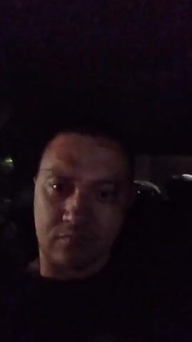 The video shows a close-up, slightly grainy, dimly lit selfie video of a person's face. The person appears to be in a car at night, as indicated by the dark environment and the slight reflection of lights in the background which suggest it could be a street or a building. The person is looking directly at the camera with a neutral expression. There is no discernable action or interaction taking place, other than the slight movement of the camera. The focus is primarily on the person's face and upper torso, which are largely obscured by darkness.