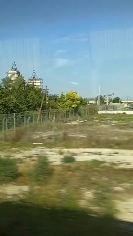 The video shows a view from inside a vehicle moving along a path. To the left, there are trees and dry grass. A fence with a sign is visible on the right side of the path. Further along the path, a white industrial building with pipes and structures on top appears among the trees. The sky is blue with some faint reflections on the window.