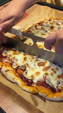 Two hands are visible slicing a long pizza with a rocking pizza cutter. The pizza has a white cheese topping and appears to have pepperoni and mushrooms. Another pizza is visible in the background, also topped with cheese and pepperoni. The pizzas are on brown parchment paper on a wooden surface. The scene is likely a kitchen.