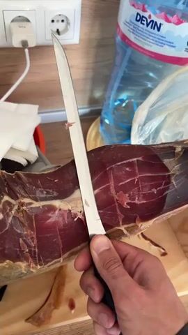 A person is shown carefully slicing a thin piece of jamón or similar cured ham from a large ham leg, which is secured in a wooden holder. The person is using a long, thin-bladed knife with a black handle. The ham appears deep red with streaks of fat. Some slices and shavings of ham are already on the wooden surface of the holder. In the background, there is a wooden countertop, a wall with an electrical outlet, a plastic water bottle, and a plastic bag. The lighting appears to be natural indoor light.