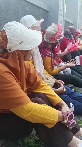 This video shows a group of people, mostly women, sitting on the ground outdoors. They are all wearing casual clothing, and many are wearing headscarves. Some of them are wearing hats. The people are sitting in a line along a wall, and some of them are looking at something in front of them, possibly a performance or an event. There are several bags and other personal items scattered around them, as well as some small flags. The environment appears to be an outdoor gathering space, possibly a park or a public square, with some trees and a building in the background. The overall mood seems to be relaxed and casual, with people waiting for something to happen.