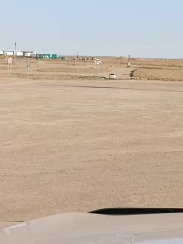 A wide shot shows a flat, dusty, open area with some sparse vegetation in the distance. Several small structures and a few signs are visible in the background. A white vehicle can be seen driving on a dirt road in the middle ground. The foreground shows the edge of a light-colored vehicle. The sky is clear and blue.