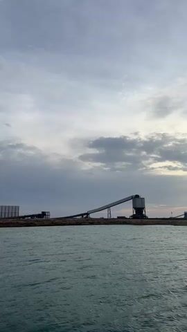 The video captures a serene waterscape at what appears to be sunrise or sunset, with a vibrant sun partially obscured by clouds. In the foreground, the water shimmers with golden reflections. The background reveals industrial structures, possibly a port or a processing plant, featuring conveyor belts, silos, and a large ship docked. The camera pans slowly from left to right, showcasing the expansive scene.