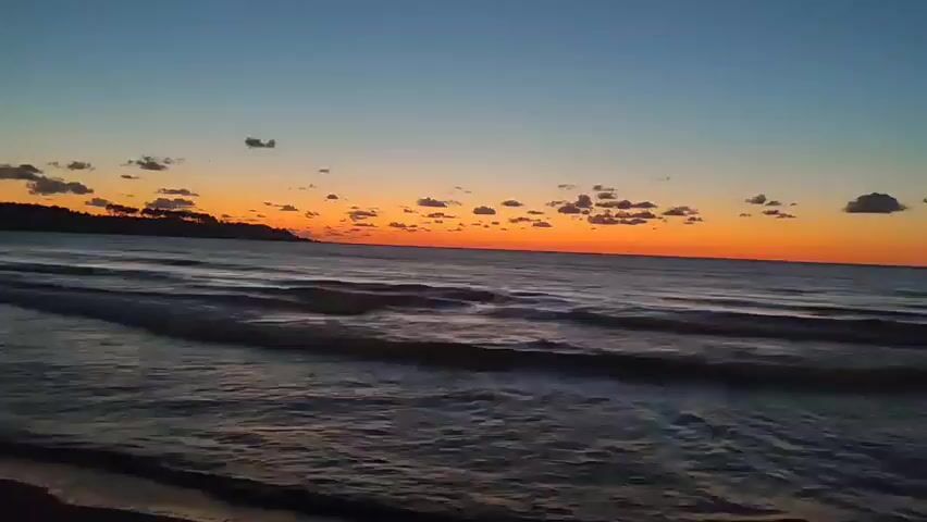 The video shows a sunset over a body of water. The sky is a gradient of blue to orange. The sun is low on the horizon, casting a warm glow. Small clouds are scattered across the sky. The water is calm, with gentle waves lapping at the shore. A silhouetted landmass is visible in the distance on the left side of the frame. The shore is dark and appears to be sandy. The overall mood is peaceful and serene.