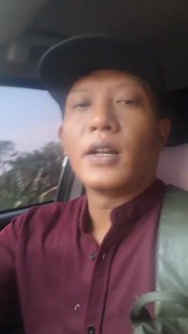The video shows a close-up of a person wearing a black baseball cap and a maroon shirt, speaking to the camera while inside a car. The person appears to be driving, with trees and the sky visible through the car window in the background. The person blinks and turns their head slightly as they speak.