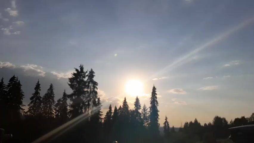 The video captures a serene outdoor scene during what appears to be sunset or sunrise. The sun, a bright orb, is positioned in the upper center of the frame, casting a strong glow and creating visible light rays that stretch across the sky. The sky is a mix of blue and white, with scattered clouds that are illuminated by the sun's rays. A silhouette of a dense line of evergreen trees runs across the lower portion of the frame, forming a natural horizon. The trees are dark and distinct against the brighter sky. The overall environment suggests a natural, possibly forested, landscape. There are no people or animals visible in the video.