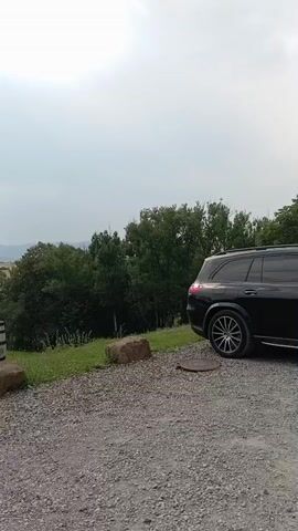 A black SUV is parked on a gravel lot. The lot is next to a grassy hill covered in rows of small trees. A wooden barrel and two rocks are in the foreground. The sky is overcast.