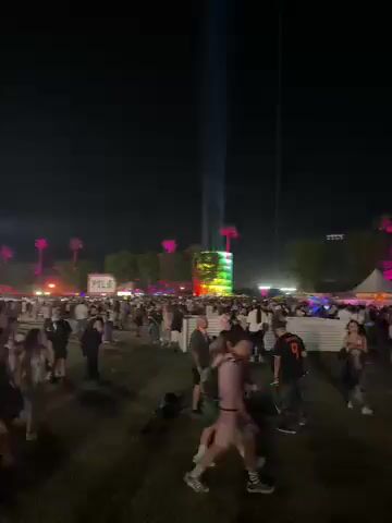 The video captures a chaotic nighttime scene at a large outdoor event, likely a music festival. A dense crowd of people occupies a vast open area, with individuals walking, standing, and moving about. In the background, multiple large structures are illuminated with bright, colorful lights; one tall structure emits a powerful beam of light into the dark sky, while another displays a vibrant, rainbow-like pattern. Portable floodlights and other artificial lights illuminate parts of the ground and the crowd. The ground appears to be an uneven, dark surface, possibly dirt or grass.

The activity primarily involves people navigating the crowded space. Notably, the video shows two instances of individuals falling or lying on the ground. At one point, a person is seen collapsing, and later, another individual is lying on their back, seemingly passed out or resting, with a small can visible near them. The overall impression is one of a lively, possibly overwhelming, festival environment. The time of day is clearly night, with no natural light visible. No specific weather conditions are discernible, other than it being an outdoor event. No visible text is present in the video. The location context of Indio, United States, strongly suggests a large-scale music festival.