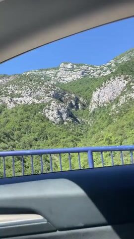 The video shows a view from inside a car of a mountainous landscape. The mountains are covered in green vegetation, with some rocky outcrops visible. A blue railing runs along the side of the road, suggesting the car is on a bridge or elevated roadway. The sky is clear and blue. The time stamp on the video indicates it was recorded on May 1st at 00:00:00. The audio appears to be music.