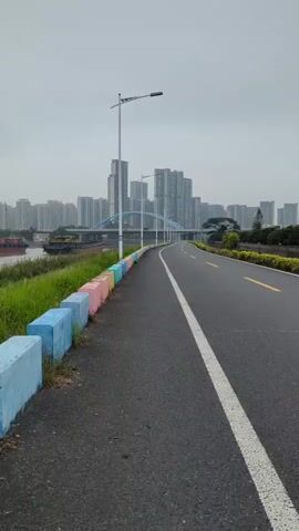 The video shows a black asphalt road curving towards a city skyline in the distance. The road has a solid white line dividing lanes and a yellow line on the right edge. On the left side of the road is a raised embankment with green grass. A row of concrete blocks lines the edge of the road, each painted a different pastel color (pink, yellow, turquoise, blue). Tall, modern buildings dominate the skyline, partially obscured by a hazy, overcast sky. A large arched bridge spans across the water in front of the buildings. To the left of the road and grass, a wide body of water has a barge moving along its surface. Tall, white lampposts line the road at regular intervals. The camera appears to be stationary, capturing the scene from a low angle. There are no people visible.