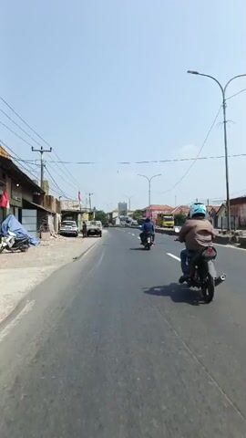 This is a first-person perspective video taken from a moving vehicle, likely a motorcycle, on a paved road. The weather is clear and sunny with a bright blue sky. The road has two lanes, separated by a white dashed line on the right side of the frame. On the left side of the road, there are buildings, some appearing to be commercial or industrial, with various signs and structures. There are also utility poles with numerous wires running overhead. Several vehicles are visible on the road, including at least two motorcycles with riders, and a few cars and trucks parked on the side of the road. The environment appears to be a suburban or semi-rural area with buildings and vegetation. The video captures the continuous movement of the vehicle along the road.