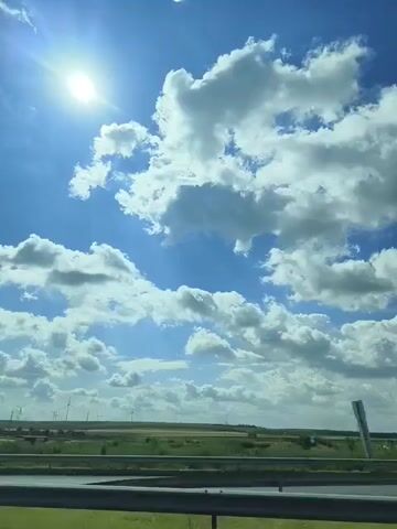 The video shows a view from a moving vehicle. The sky is blue with scattered white clouds, and the sun is shining brightly. In the distance, there is a flat landscape with fields and some construction or industrial elements, including what appear to be power lines and possibly wind turbines. The road or surface the vehicle is on is visible in the foreground, with a barrier or railing. There are no people or discernible animals in the video. The overall impression is of a daytime journey through a rural or semi-industrial area.