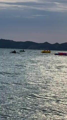 The video shows a wide shot of a body of water under a cloudy sky. In the distance, a hilly coastline is visible. Scattered across the water are several recreational watercraft. Closer to the foreground, there are two kayaks with people in them, one of which is moving. To the right of the kayaks, a yellow, car-shaped inflatable boat is visible. Further to the right, there are some red inflatable objects. On the left side of the frame, a large, pink inflatable object shaped like a cartoon character is present. A jet ski is seen moving between the kayaks and the yellow boat, and appears to be spraying water. Another jet ski is visible further in the distance. The water is moderately choppy. The overall impression is a recreational scene on the water with various water toys and people enjoying activities.