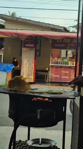 The video shows an outdoor food stall. In the foreground, a large, flat cooking surface is heated by flames from below, and several long, cylindrical food items are being fried. Behind the cooking area, a red telephone booth stands near a food stand with the sign "ES TEH JU." A person wearing a dark t-shirt and cap is seated to the left, looking at their phone. The overall scene appears to be in a warm, possibly tropical, climate, with natural light suggesting either early morning or late afternoon.
