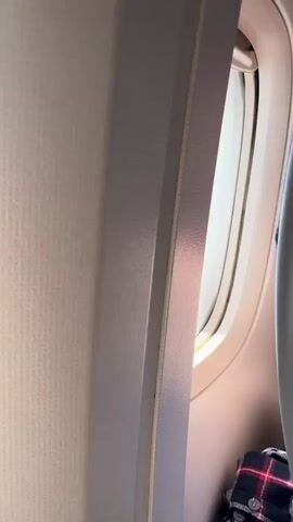 The video shows the interior of an airplane cabin with a view out of a window. The first few seconds focus on the window frame and the textured interior paneling of the airplane. Then, the view transitions to the outside of the airplane, showing the wing and a vast expanse of white, fluffy clouds stretching out below. The sky is visible above the clouds, appearing bright and hazy with some sunlight glinting off the wing. There are no people or other objects clearly visible outside the airplane. The video appears to be a static shot from the perspective of a passenger looking out the window during a flight.