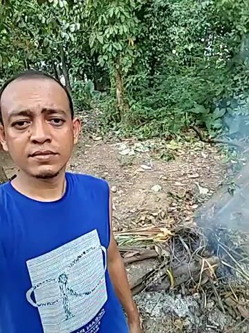 A man is standing in a forest-like setting. He has a shaved head and is wearing a blue sleeveless shirt. He's looking directly at the camera, which he is holding. Behind him, there is a small fire with smoke rising from it. There are trees and foliage visible in the background. The ground is covered with leaves and fallen branches.