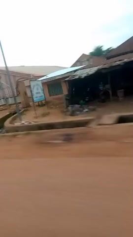 The video shows a side view of a dirt road with some buildings and trees. The camera is moving, causing a blur effect. In the foreground, there is a reddish-brown dirt road. To the left, there are some buildings with light-colored walls and roofs, and some green vegetation. In the middle ground and background, there are more buildings, some with brown walls and roofs, and some trees. The sky is overcast. There is no clear indication of people or specific actions taking place, other than the implied movement of the camera. The overall scene appears to be a residential or semi-rural area.