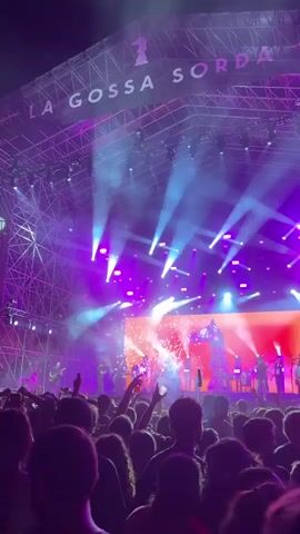 A crowd of people are watching a concert at night. A band is playing on a large stage illuminated with purple and white lights. Spark effects go off in front of the stage. The band is visible as silhouettes against a bright red screen. The name "LA GOSSA SORDA" is written across the top of the stage structure. Many people in the crowd are holding up their phones to record the concert.