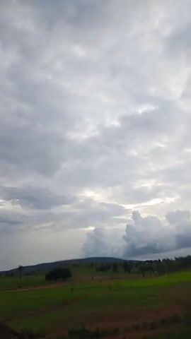 A cloudy sky looms over a green landscape. In the distance, hills can be seen under the clouds. The foreground shows a grassy field with a few trees and some barren ground. The scene appears to be a rural area, possibly a farm or open countryside. The lighting suggests it might be overcast or close to sunset/sunrise. No people or specific actions are visible.