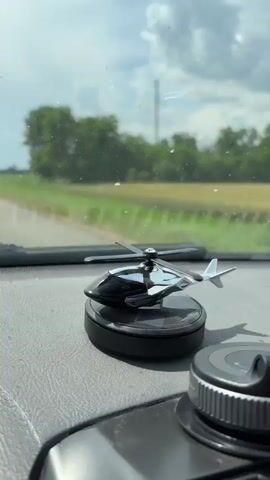 A close-up shot shows a silver helicopter toy with spinning blades perched on a black circular base. The toy is positioned on a gray dashboard inside a vehicle. In the background, through the windshield, a rural landscape is visible, featuring a road, green fields, and trees under a cloudy sky. The shot is slightly shaky, suggesting the vehicle is in motion.