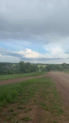 The video shows a dirt road cutting through a grassy field under a cloudy sky. In the first frame, a collection of trees stands near a pond, with buildings visible in the distance. The following shots show the road winding through the landscape, with trees lining the edges of the field. The sky transitions from partly cloudy with patches of blue to a more overcast, grey sky. The overall scene is rural and undeveloped.