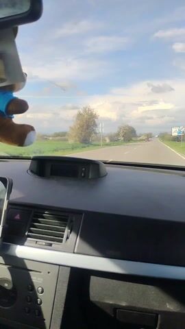 The video shows a view from inside a car driving on a two-lane road. The sky is blue with some white clouds, and the landscape includes green fields and some trees with white blossoms. There are other cars on the road in the distance. The interior of the car is visible, including the dashboard and a side mirror showing the passing scenery. There is a small Bulgarian flag attached to the passenger side pillar of the windshield.