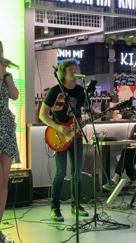This video captures a live music performance, likely in a public indoor space, possibly a shopping mall or a venue with a modern aesthetic, given the visible signage and bright lighting. The central figure is a male guitarist and singer, wearing a black t-shirt and jeans, actively playing an electric guitar that is a striking red and yellow. He is positioned behind a microphone, suggesting he is singing. To his left, a female individual is partially visible, wearing a patterned dress and also holding a microphone, implying she might be a vocalist or part of the performance.

In the background, another person is seated at a keyboard or DJ setup. The setting features neon green lighting on the left side of the frame, and various signs are visible, including text in what appears to be Cyrillic and English. One sign reads "NH MI VIETNAMESE SANDWICH", indicating a potential food establishment nearby. Another sign displays "KIA". The overall atmosphere seems energetic, with the musicians engaged in their performance. The lighting suggests it could be daytime or early evening, and there are no obvious indicators of weather conditions. The presence of multiple microphones, amplifiers (one clearly labeled "Marshall"), and musical instruments establishes the scene as a musical event. The focus is on the live performance, with the musicians appearing concentrated on their act.